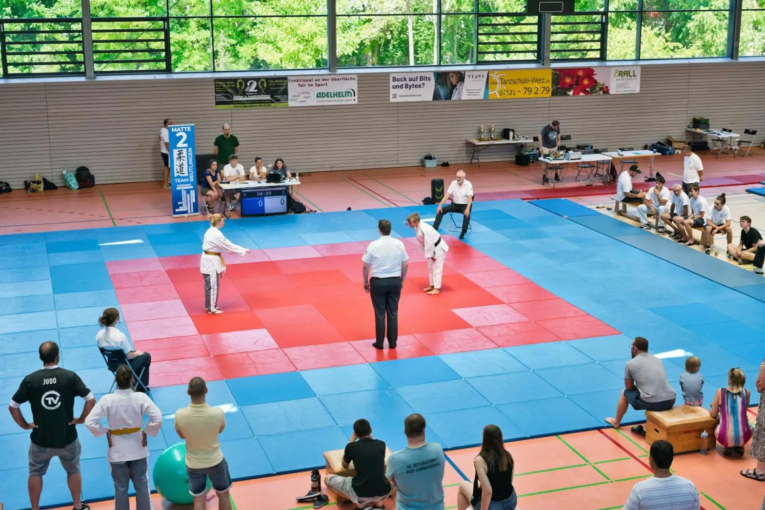Turnaj v judu v hale: dva závodníci a rozhodčí na modro-červeném tatami, kolem diváci