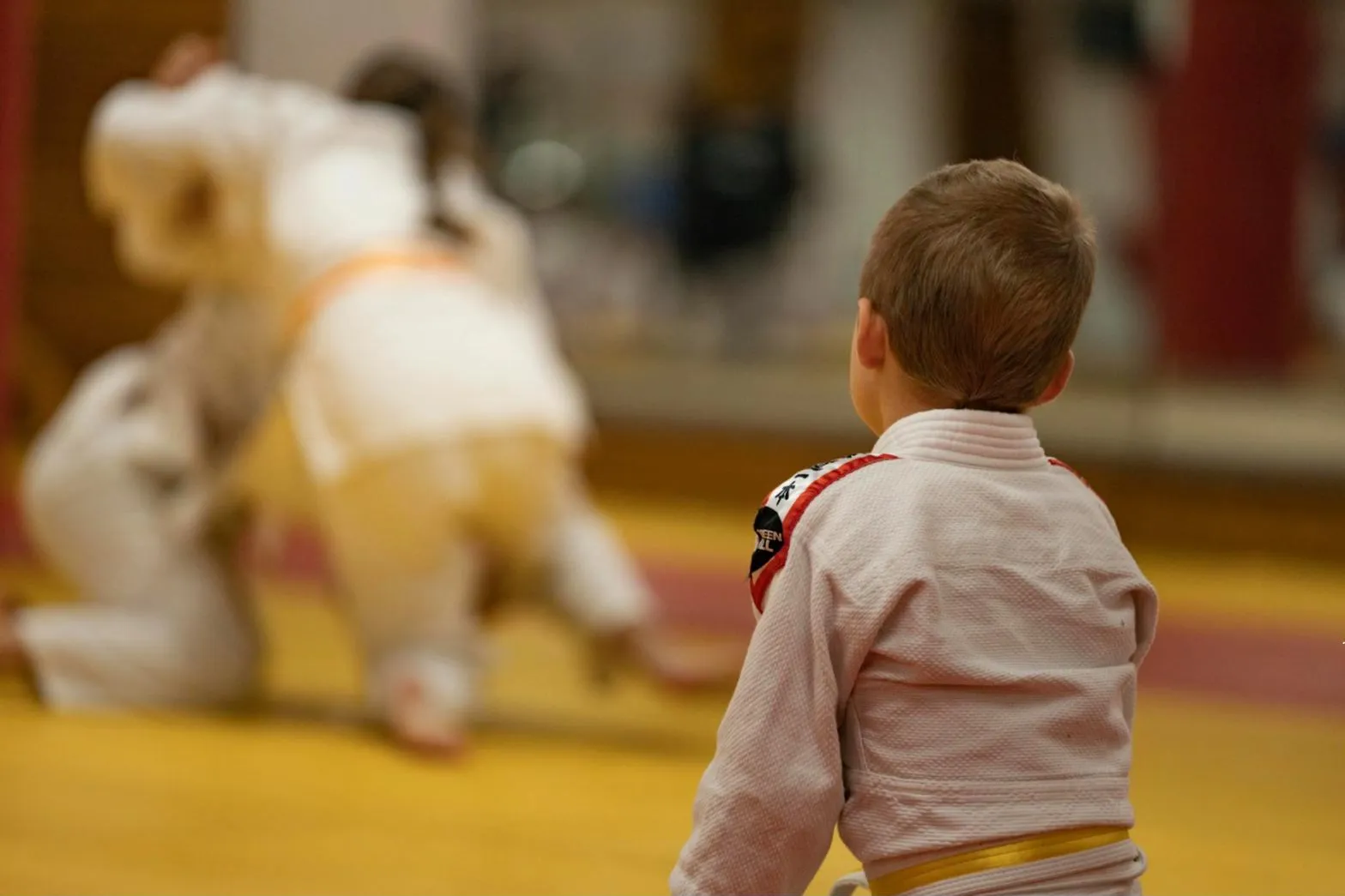 Chlapec v kimonu sleduje zápas dvou dětí na tatami